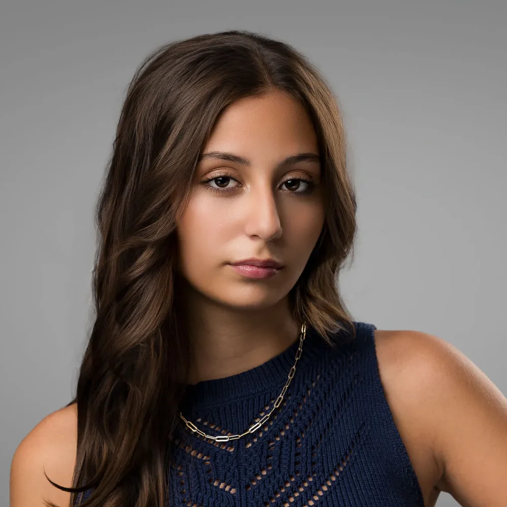 Editorial portrait of a young woman with long brown hair in a navy knit top on a gray backdrop