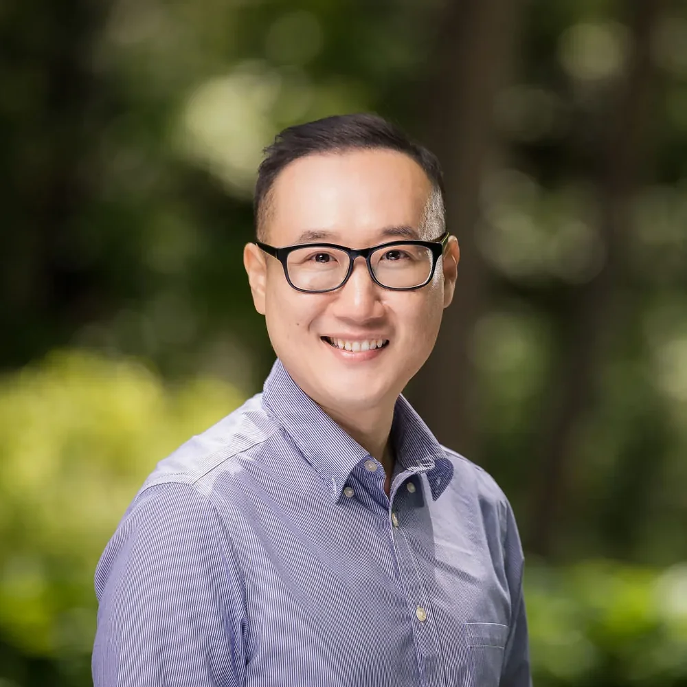 Outdoor headshot of a man in glasses and a blue striped shirt with green bokeh background