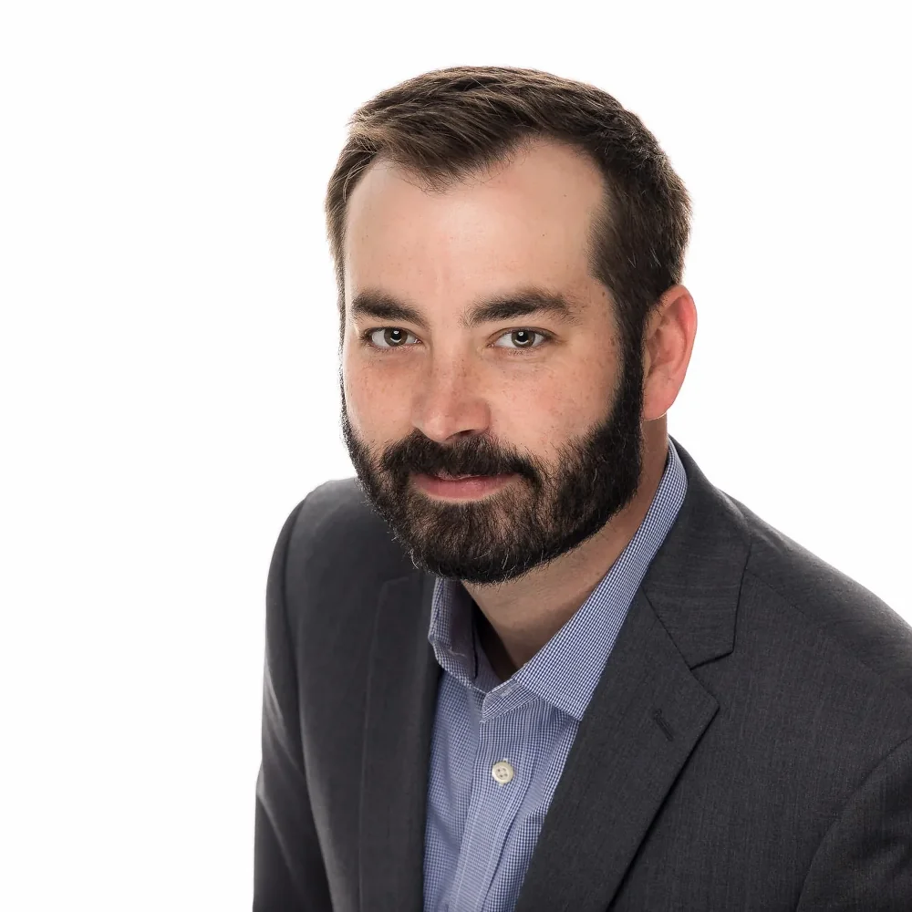 Professional headshot of a bearded man in a gray blazer over a blue checked shirt on a white backdrop