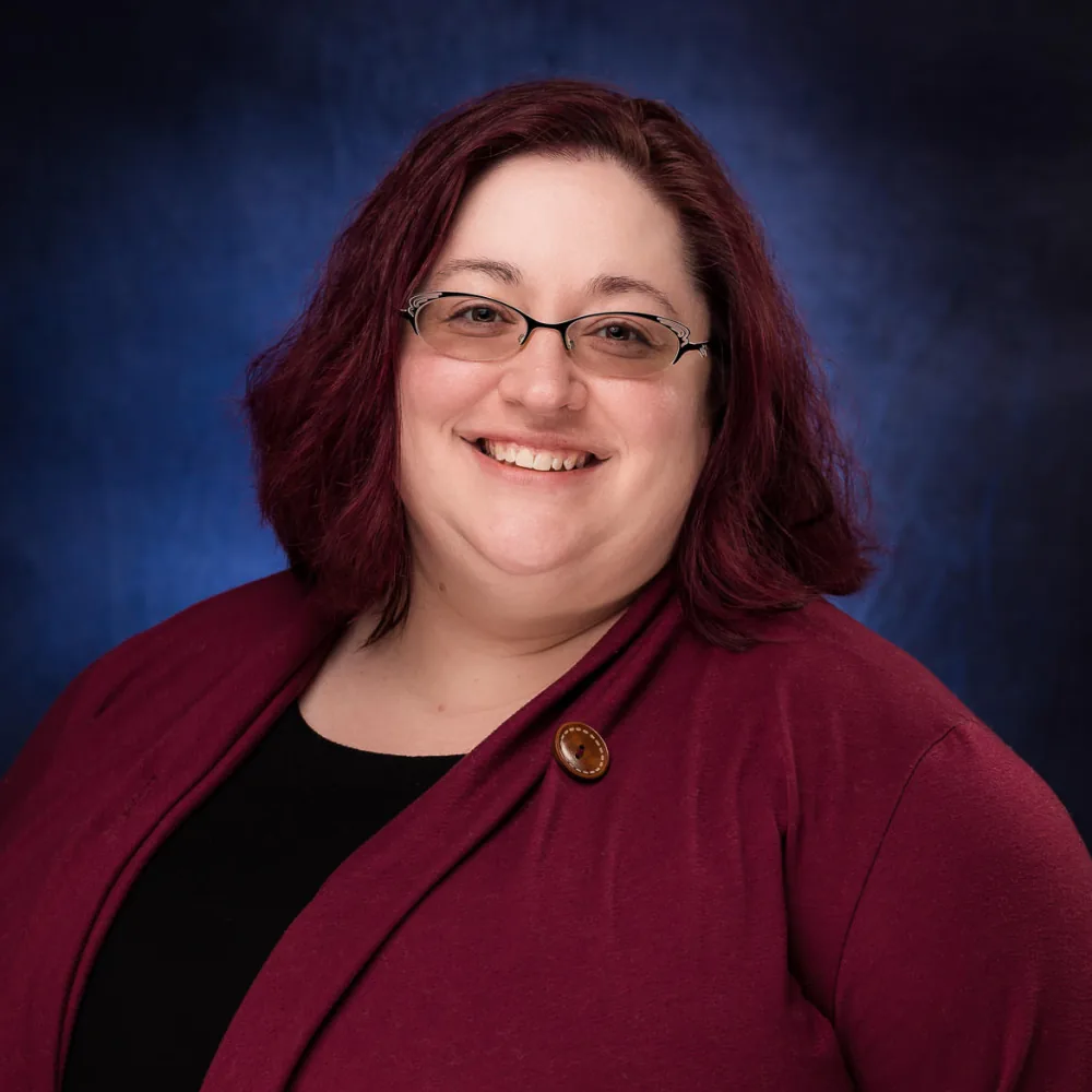Studio headshot of a woman with dark red hair and glasses in a maroon blazer on a deep blue backdrop