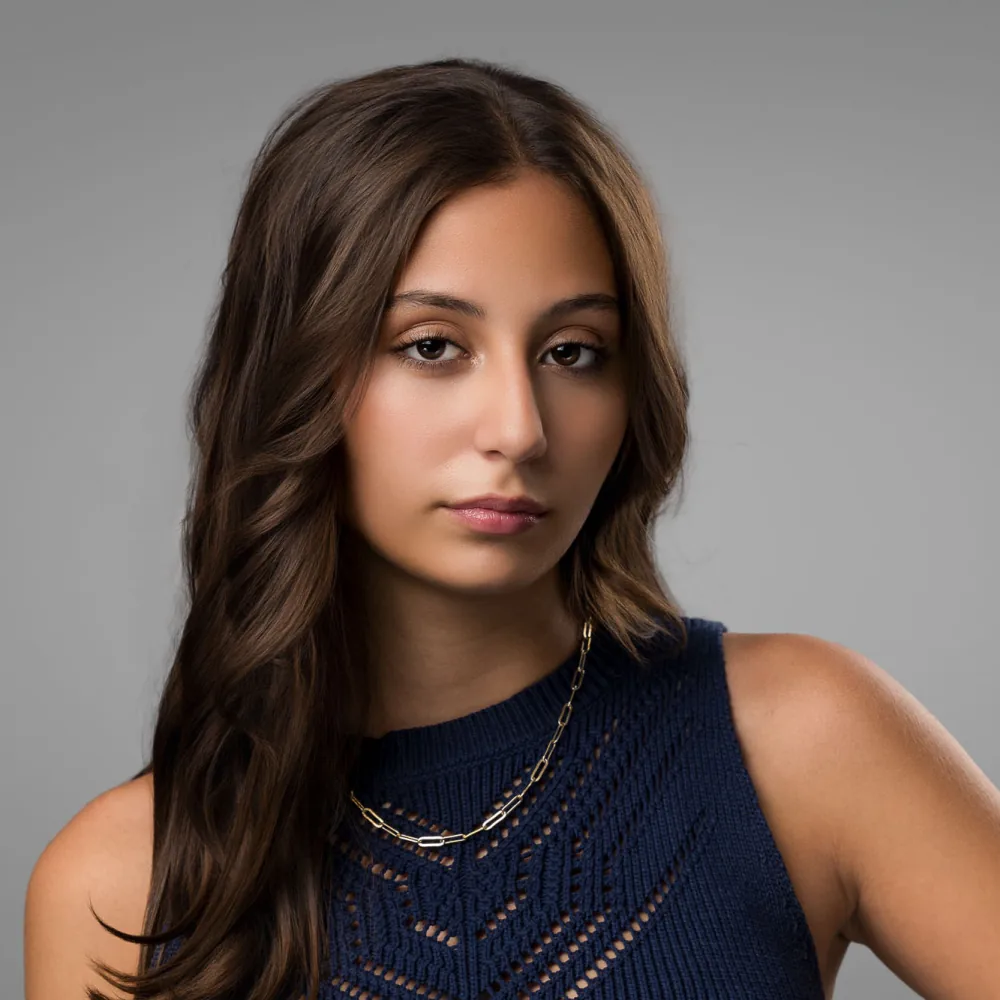 Studio headshot of a young woman with long dark hair in a navy sleeveless top on a gray backdrop