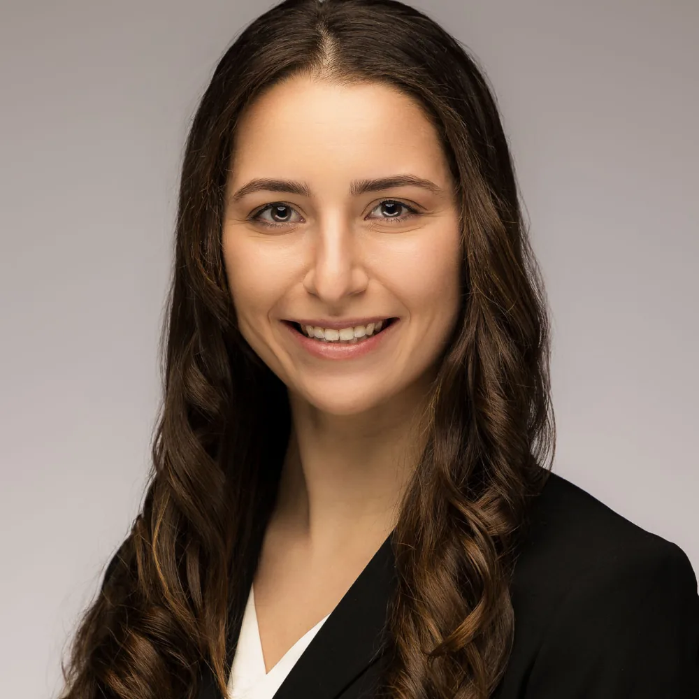 Studio headshot of a young woman with wavy brown hair in a black top on a soft gray backdrop