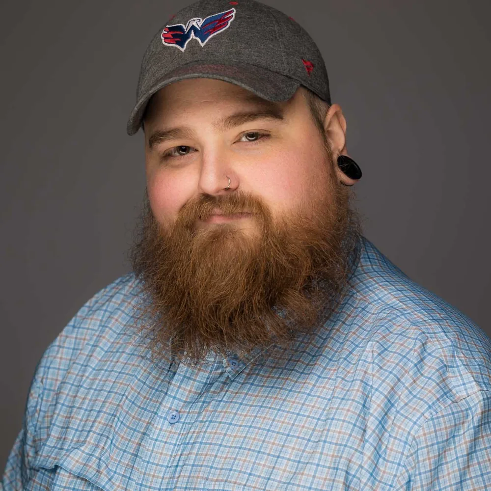 Studio headshot of a bearded man in a ball cap and blue plaid shirt on a gray backdrop