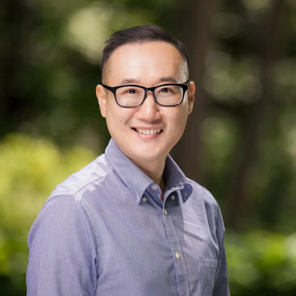 Outdoor headshot of a man in glasses and a light blue striped shirt with a green background