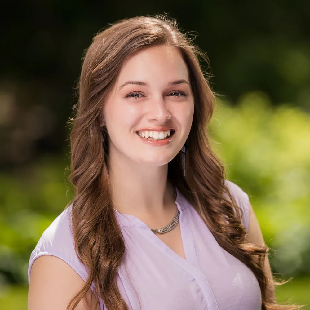 Outdoor headshot of a young woman with long brown hair in a lavender top with a green background
