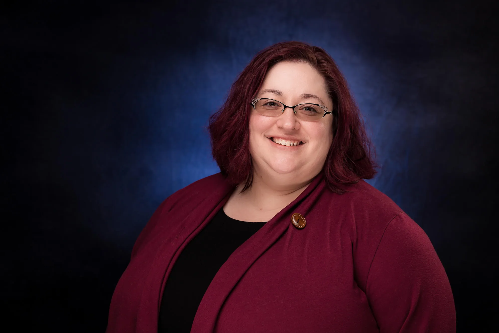 Corporate headshot of a woman in a dark red blazer on a blue mottled studio backdrop